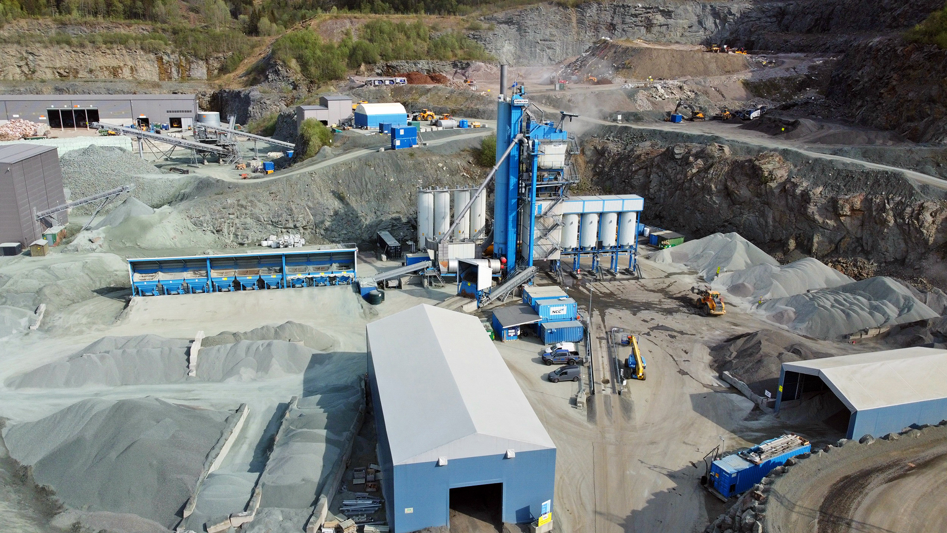 A large industrial Asphalt site with blue and white equipment for processing building materials, surrounded by piles of gravel and storage buildings.